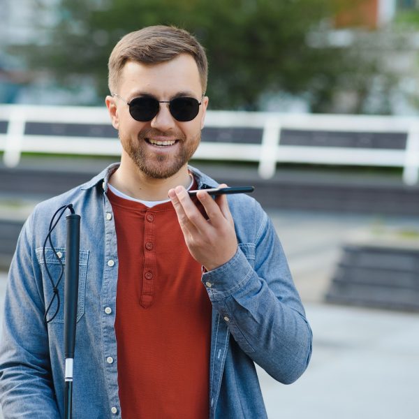 A blind man wearing sunglasses and holding a cane, speaks on the phone.