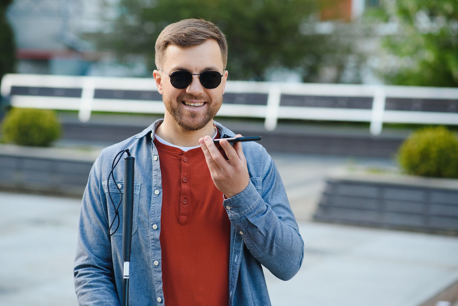 A blind man wearing sunglasses and holding a cane, speaks on the phone.