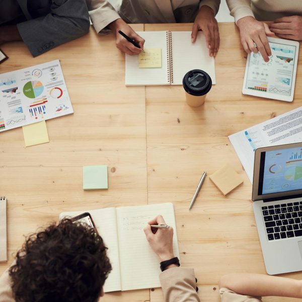 A view of a meeting where various people are taking notes on paper and on laptop, and there are printouts with charts and graphs.