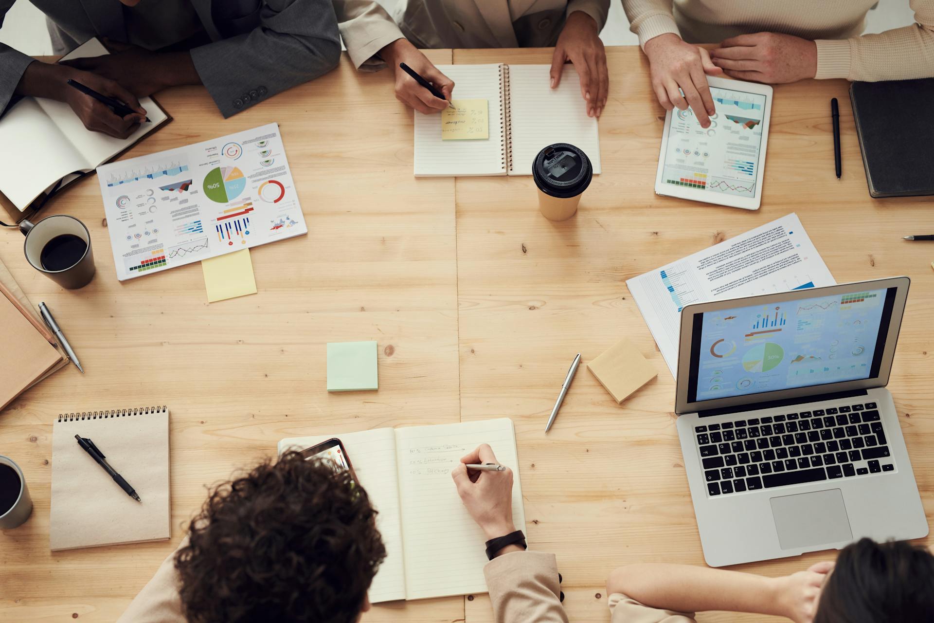 A view of a meeting where various people are taking notes on paper and on laptop, and there are printouts with charts and graphs.