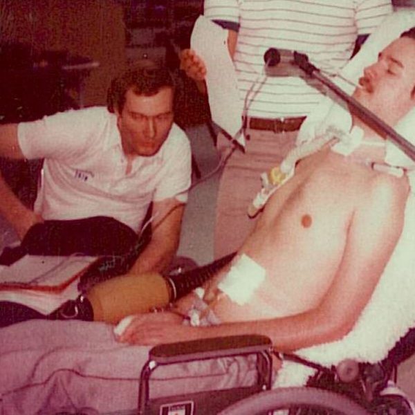 Gary Birch, in a wheelchair, looks over Neil, in a bed with a sip and puff device mounted.