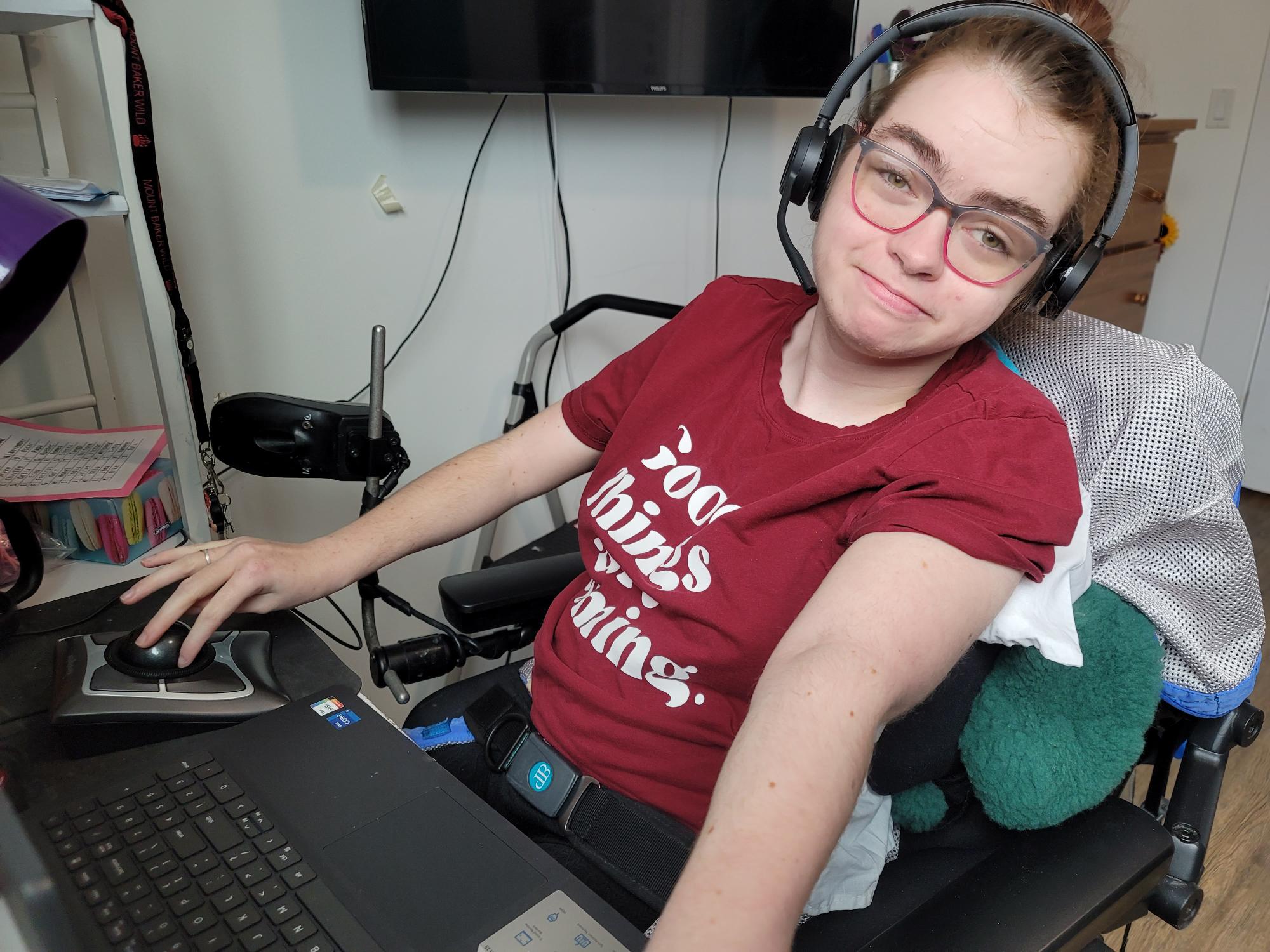 Hailey, in her wheelchair, at her desk using a trackball mouse and wearing a headset.