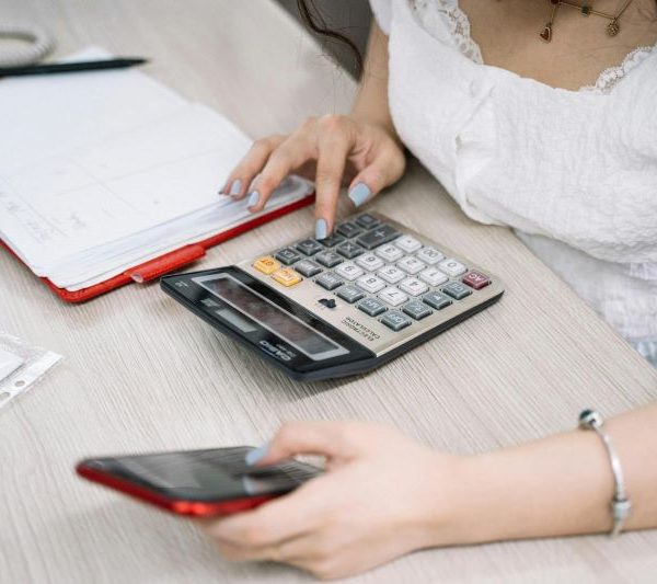 A woman holds a smartphone and uses a calculator, with some documents to the side.