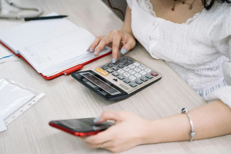 A woman holds a smartphone and uses a calculator, with some documents to the side.