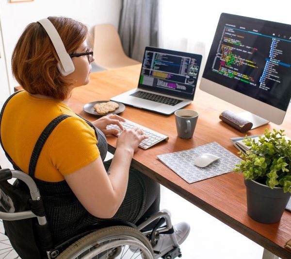 A woman in a wheelchair uses a laptop and a monitor to do some coding.