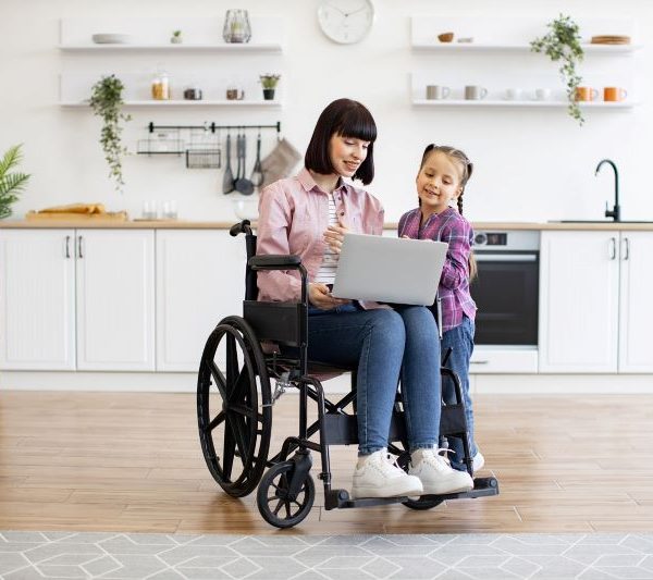 A woman in a wheelchair shows a young girl something on a laptop.