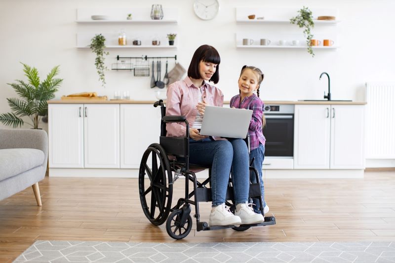 A woman in a wheelchair shows a young girl something on a laptop.