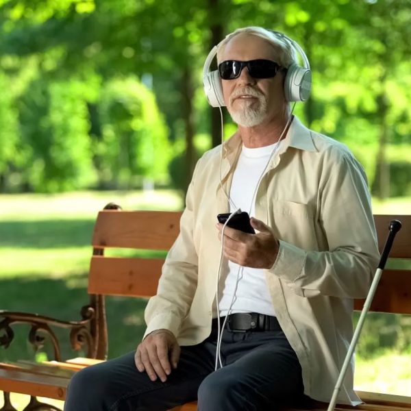 A blind man wearing sunglasses sits on bench wearing headphones and holding his phone.