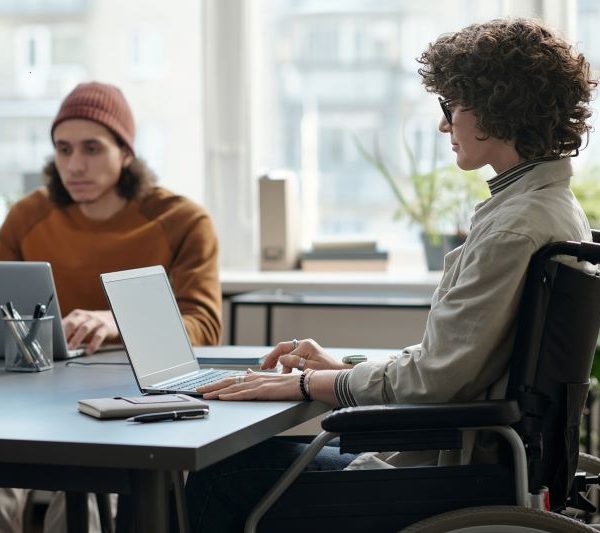 A woman in a wheelchair uses a laptop at a table, sitting close to a man also using a laptop.