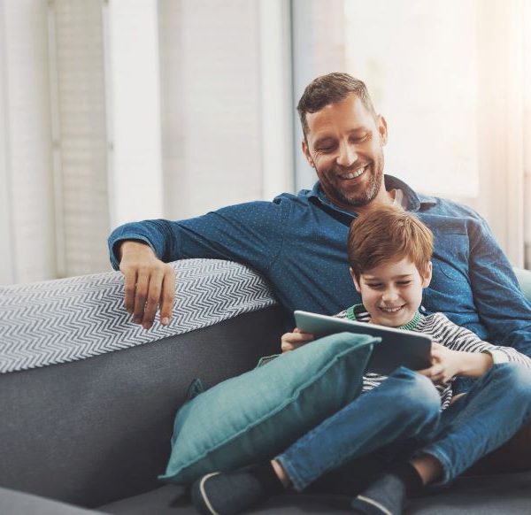 A father smiles as he watches his son use a tablet.